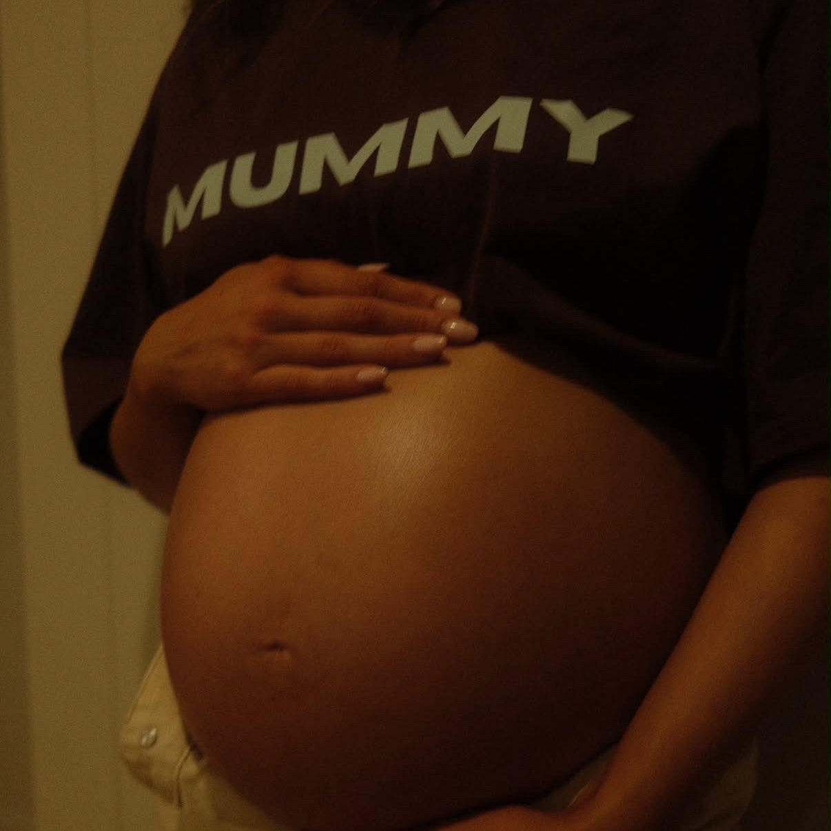 A pregnant person wearing a dark brown T-shirt with the word 'MUMMY' printed in white across the chest, holding their belly. The photo is taken in a dimly lit indoor setting.